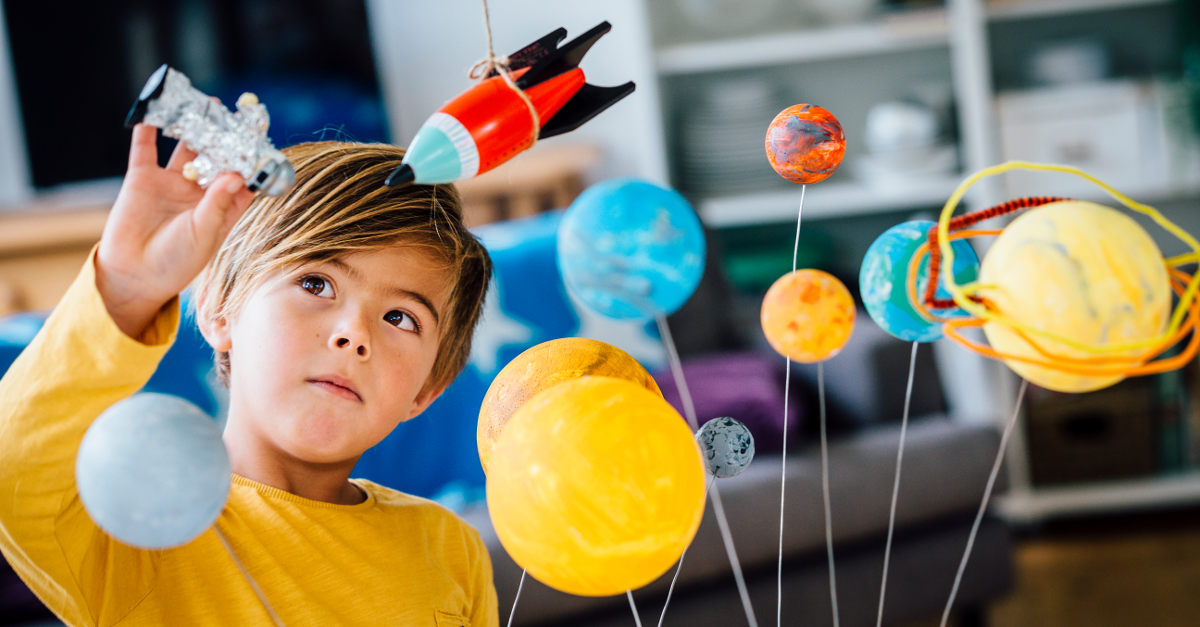 Criança brinca com astronauta de brinquedo próximo de maquete com planetas e foguete. 